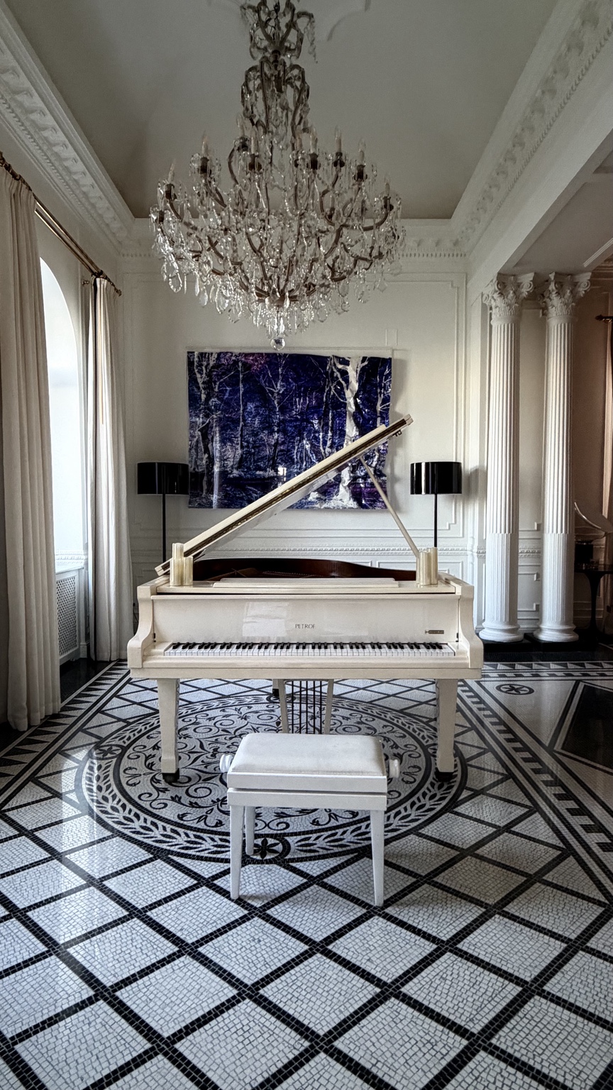 White grand piano beneath a chandelier in a Villa Bismarck room with patterned mosaic floor